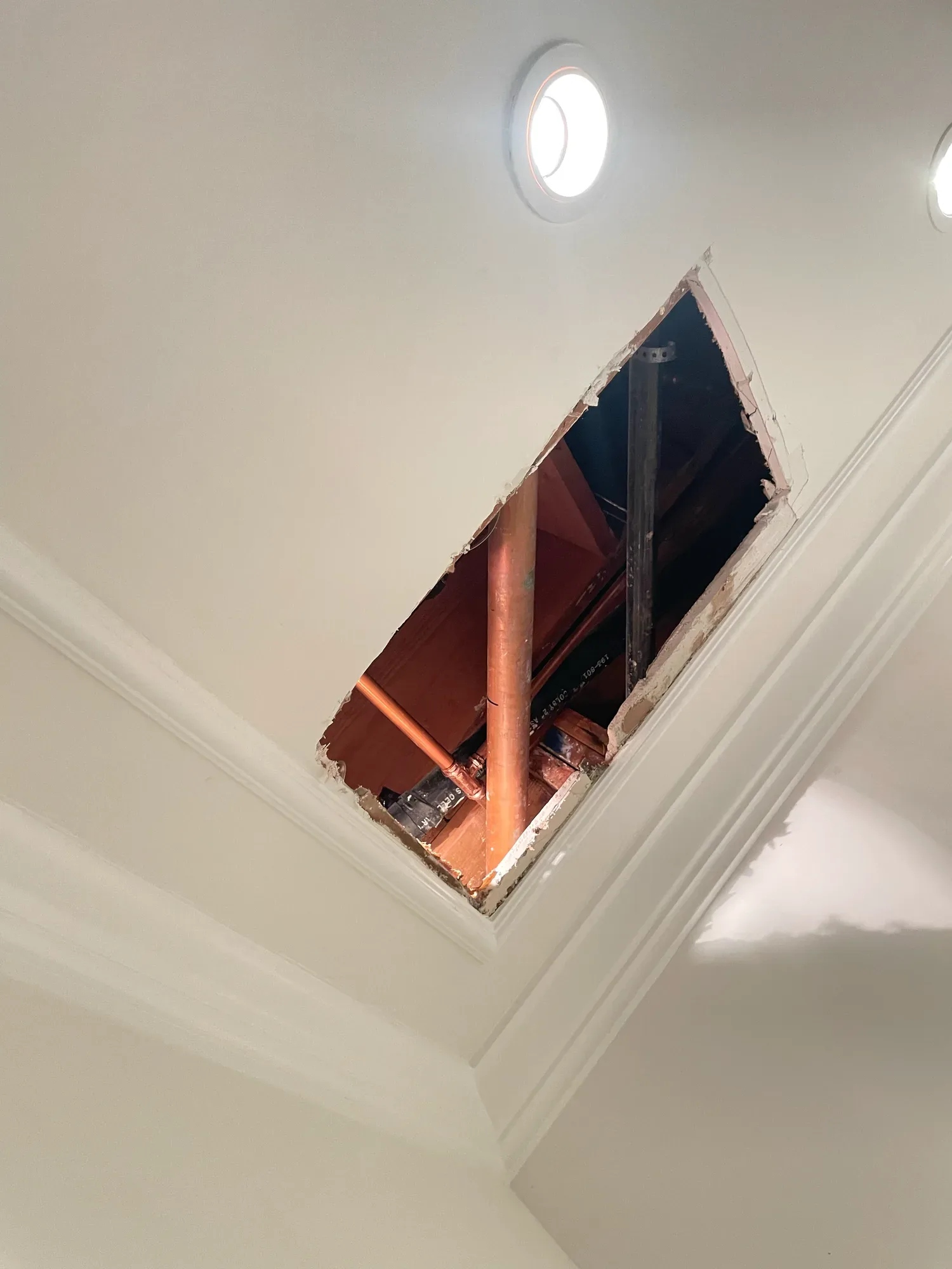 Access hole cut into a finished ceiling with crown molding, exposing a copper water pipe and a drain line