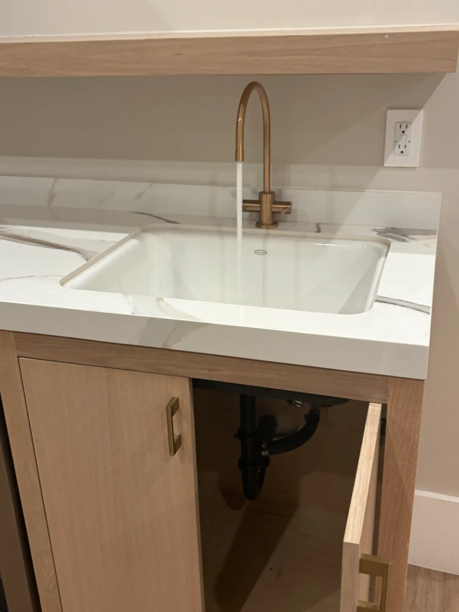 Brass faucet running into a white undermount sink, with the drain trap visible in the open cabinet below