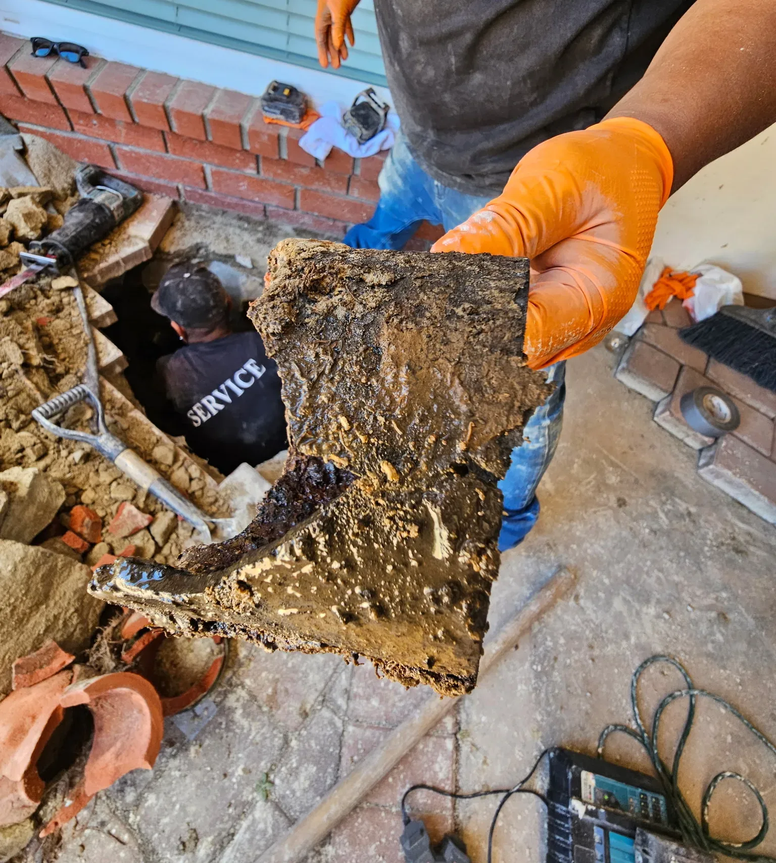 Gloved hand holding a broken section of old sewer pipe above an excavation where a technician works