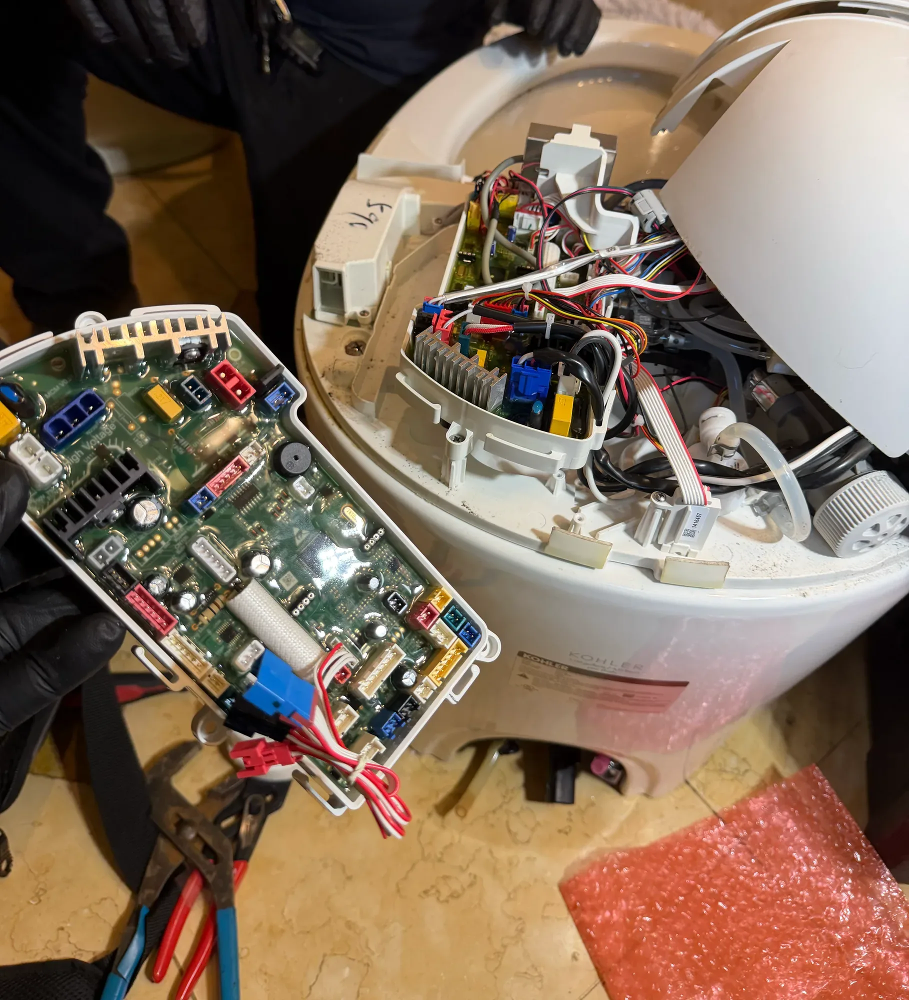 Gloved technician holding a circuit board beside an opened Kohler smart toilet