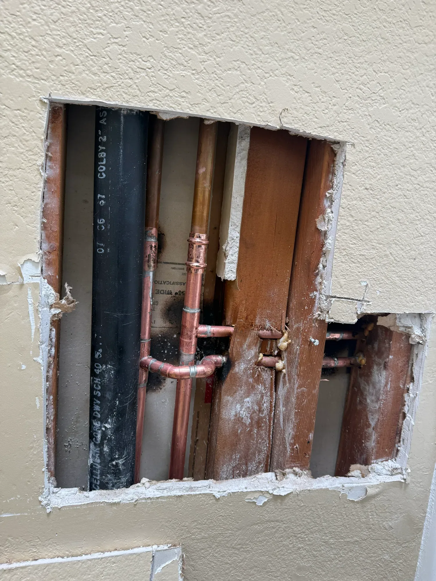 Copper water lines and fittings beside a black drain stack inside a square opening cut in a textured wall