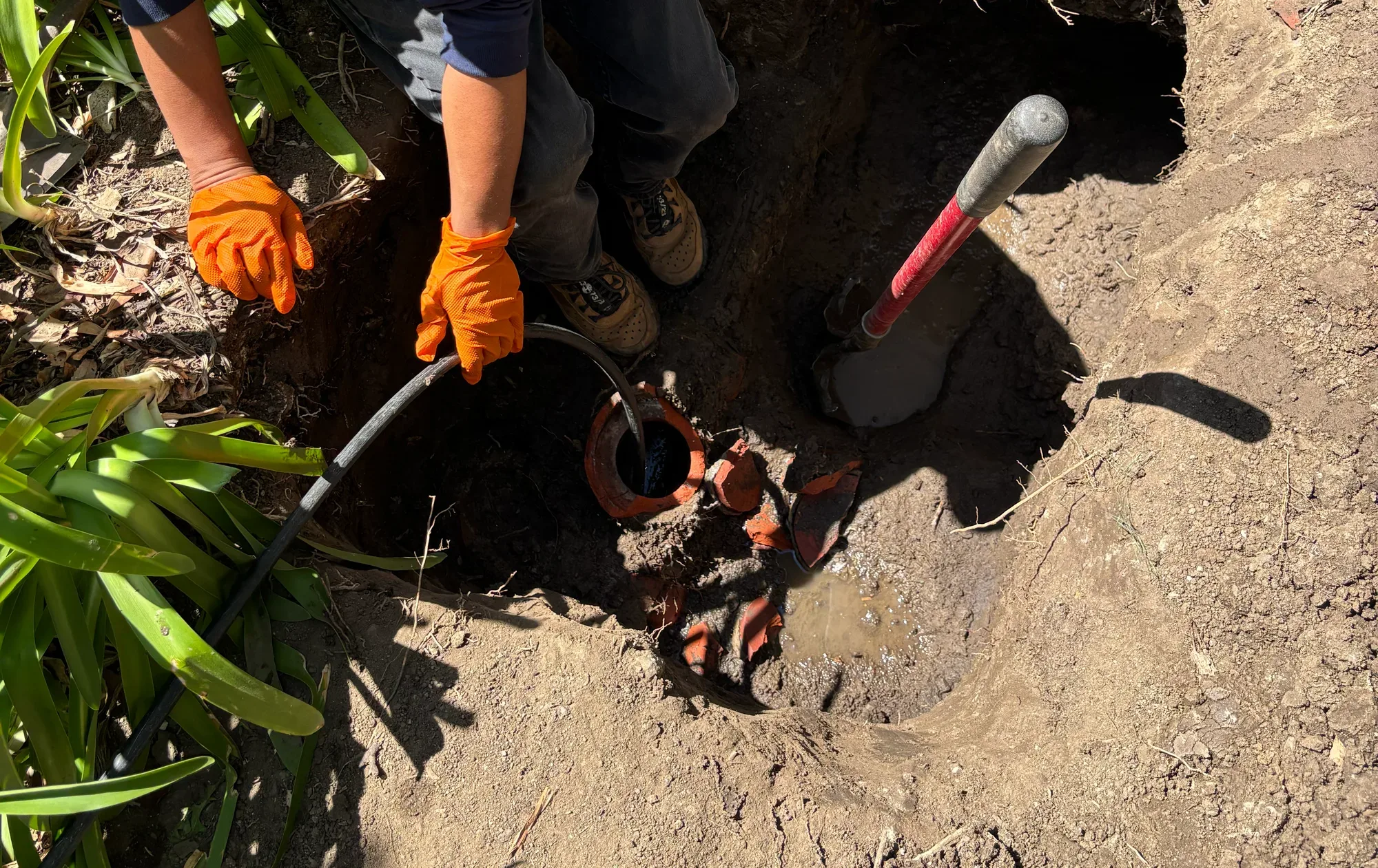 Gloved technician working at an exposed clay sewer pipe at the bottom of a dug pit, broken clay shards beside it