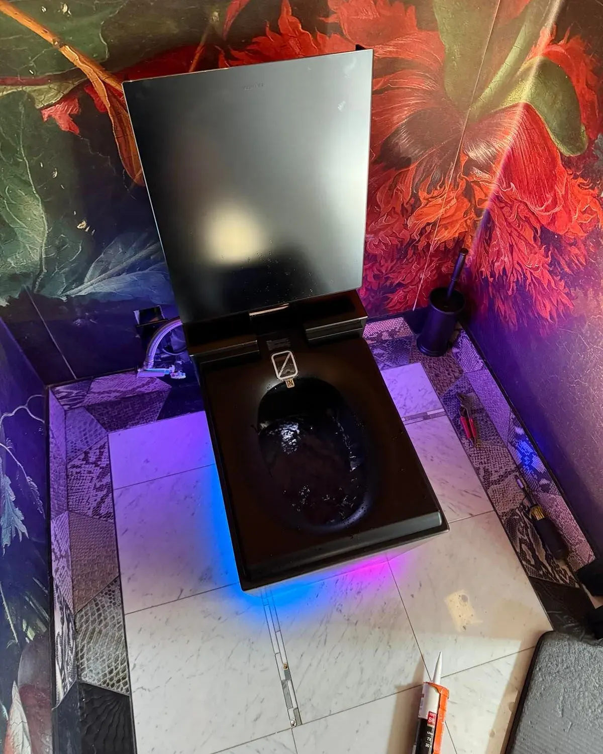 A finished Kohler intelligent toilet with the lid raised and its base lighting on, installed in a papered powder room