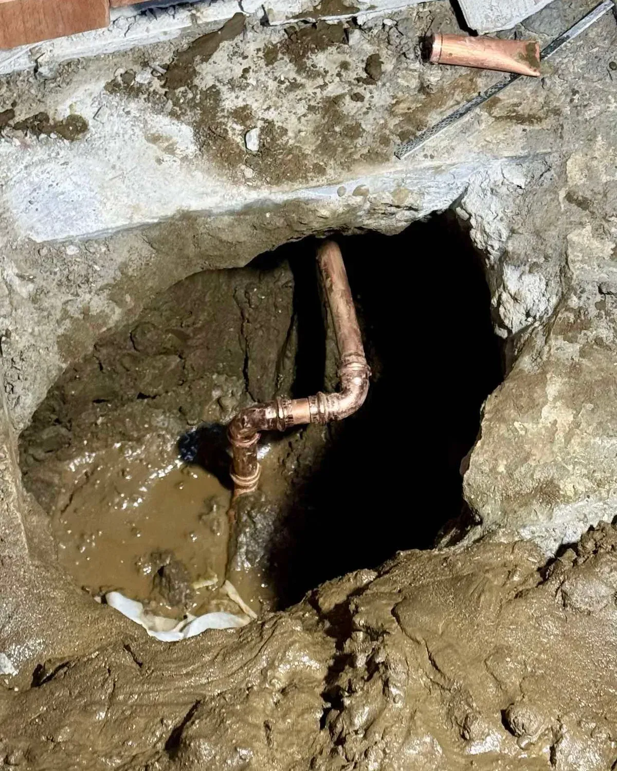 A concrete slab cut open and excavated to expose a home's copper water line, with new elbow fittings on the pipe above standing water at the bottom of the hole