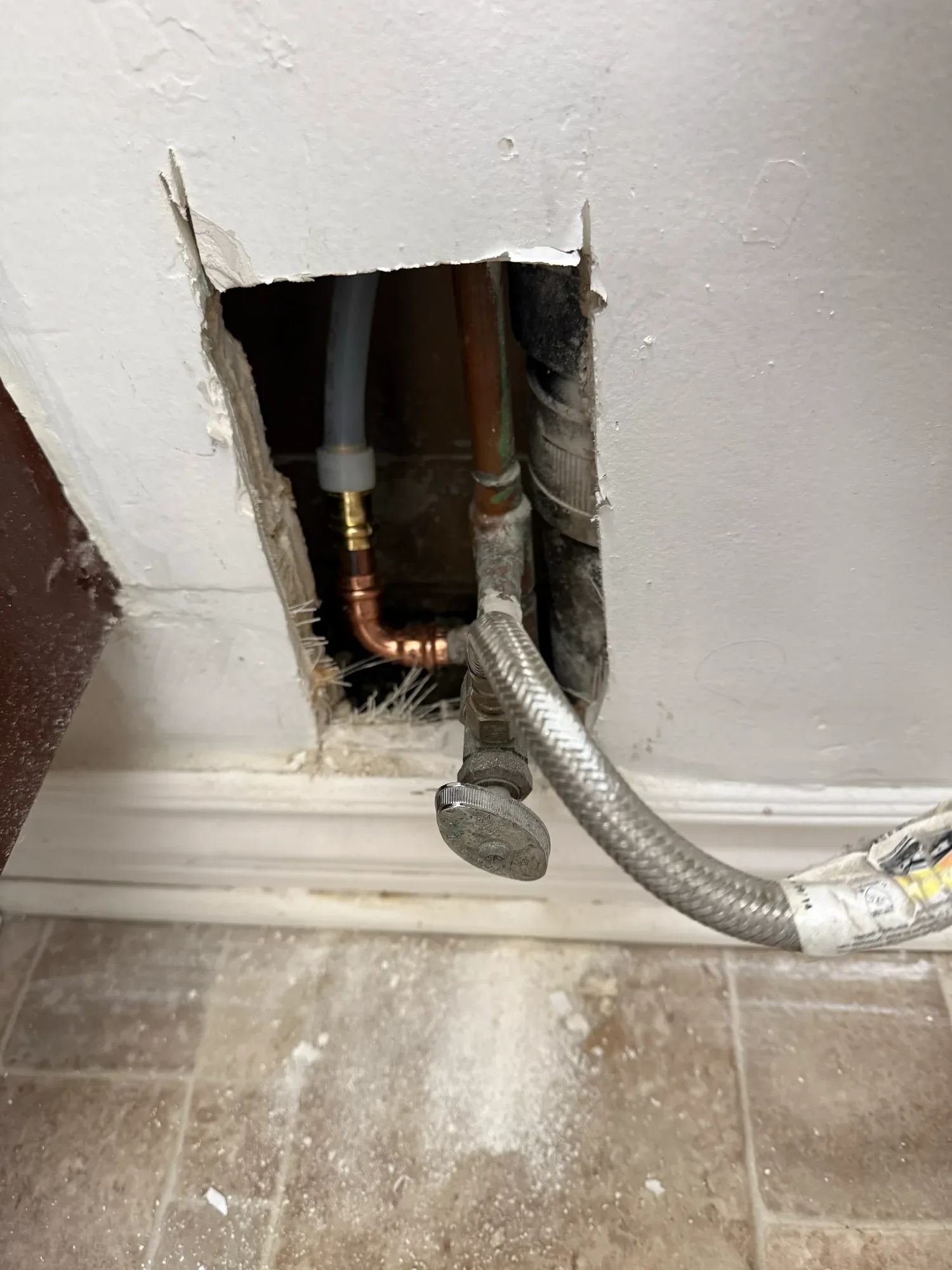 Small opening in a bathroom wall showing a new copper elbow joined to a plastic water line behind a supply hose