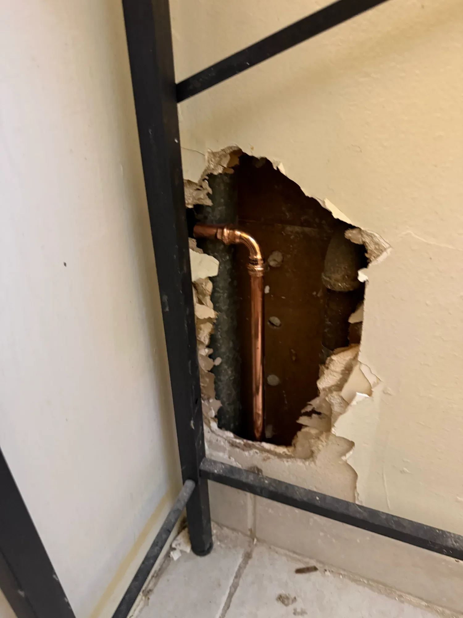 New copper elbow and line inside a rough opening in a wall, behind a black metal frame on a tiled floor