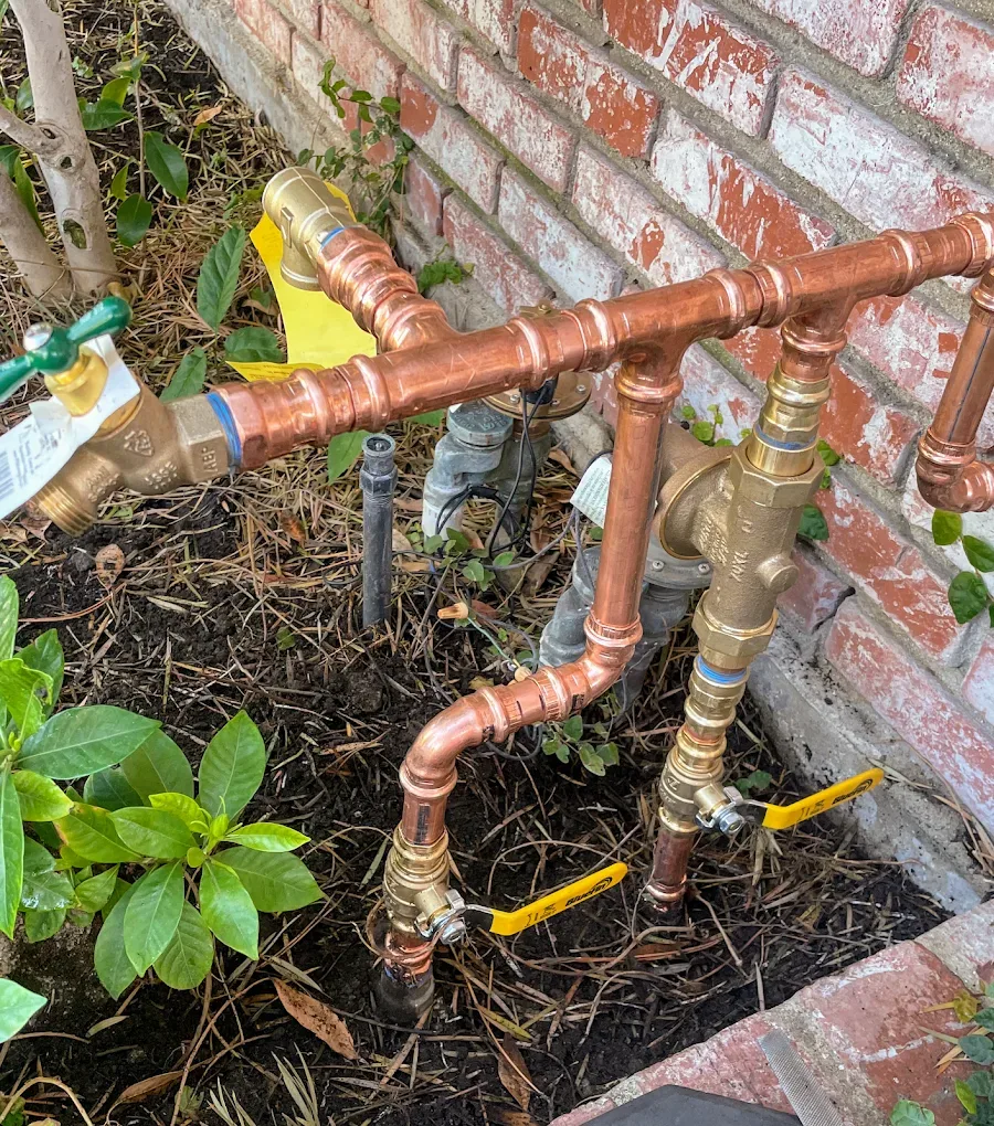 New copper water manifold with brass valves and yellow shutoff handles in a planter beside a brick wall