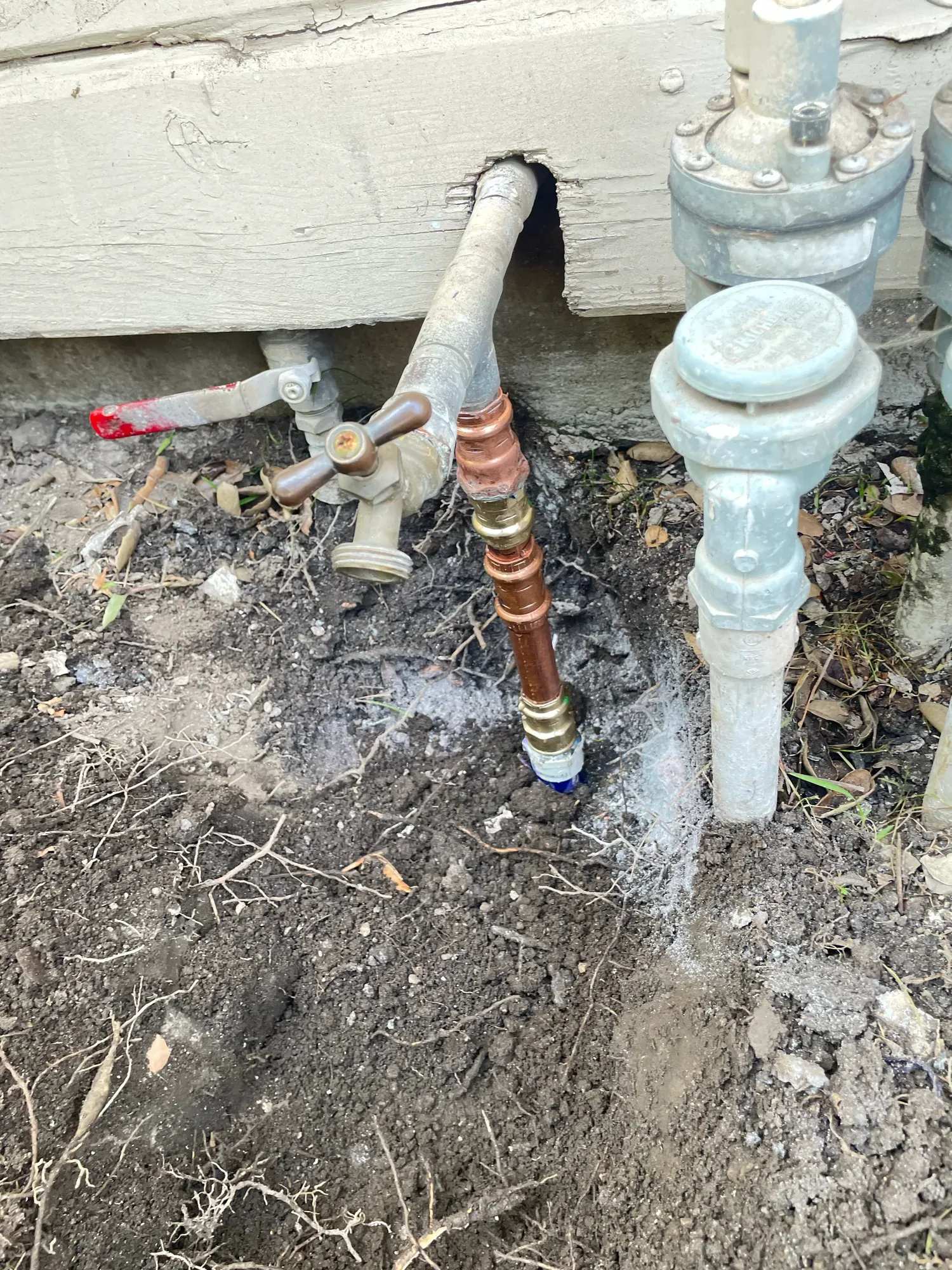 Old water line at a house wall with a new copper and brass section running into the soil, beside outdoor valves