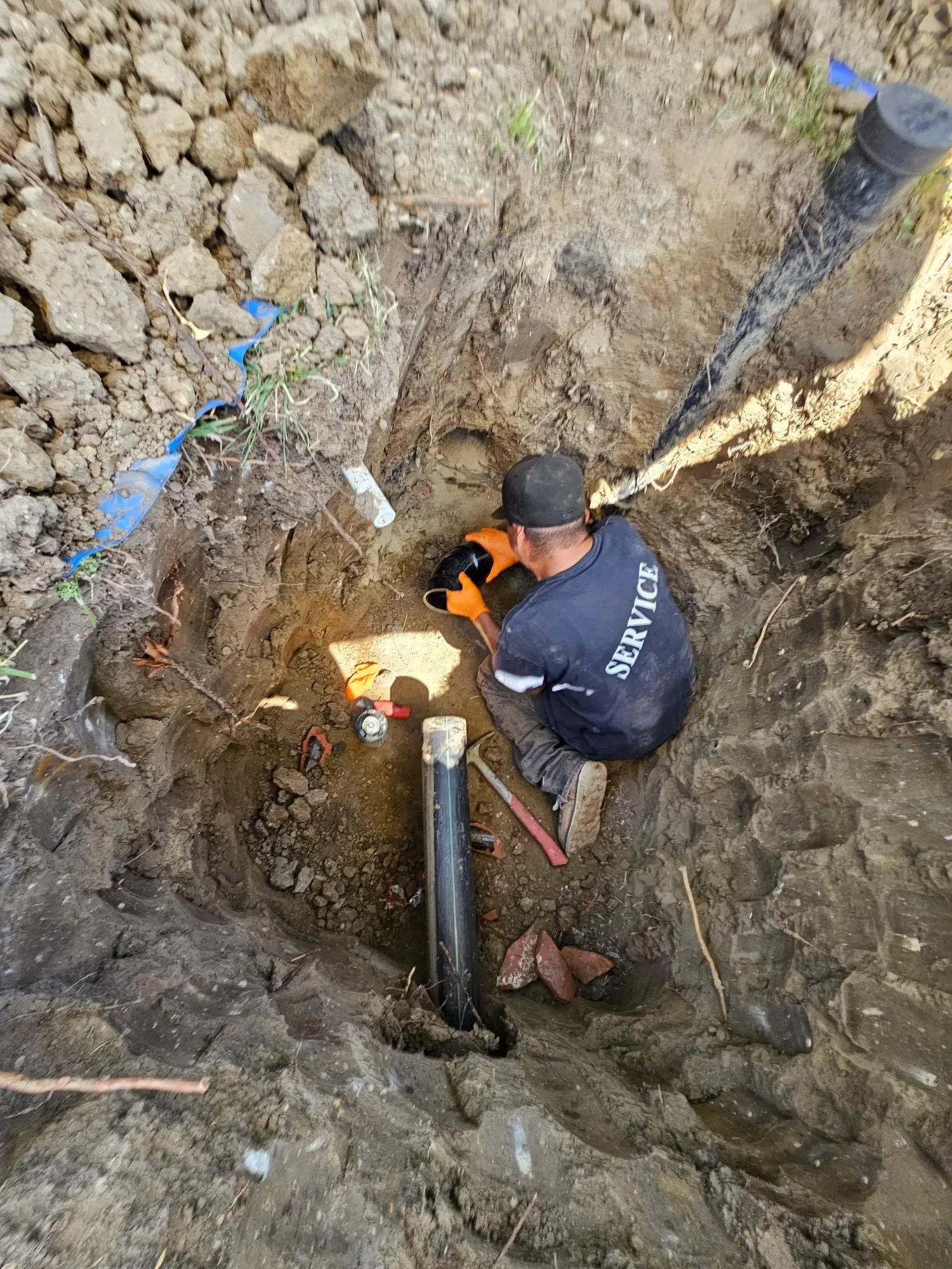 Technician in a deep excavation holding a black pipe fitting beside new pipe and broken clay pipe pieces