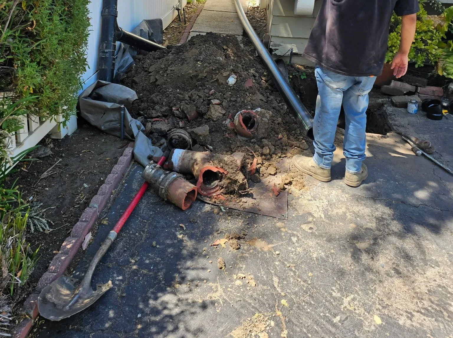 Sections of old clay sewer pipe piled on excavated soil along a side yard path, a technician at the open trench