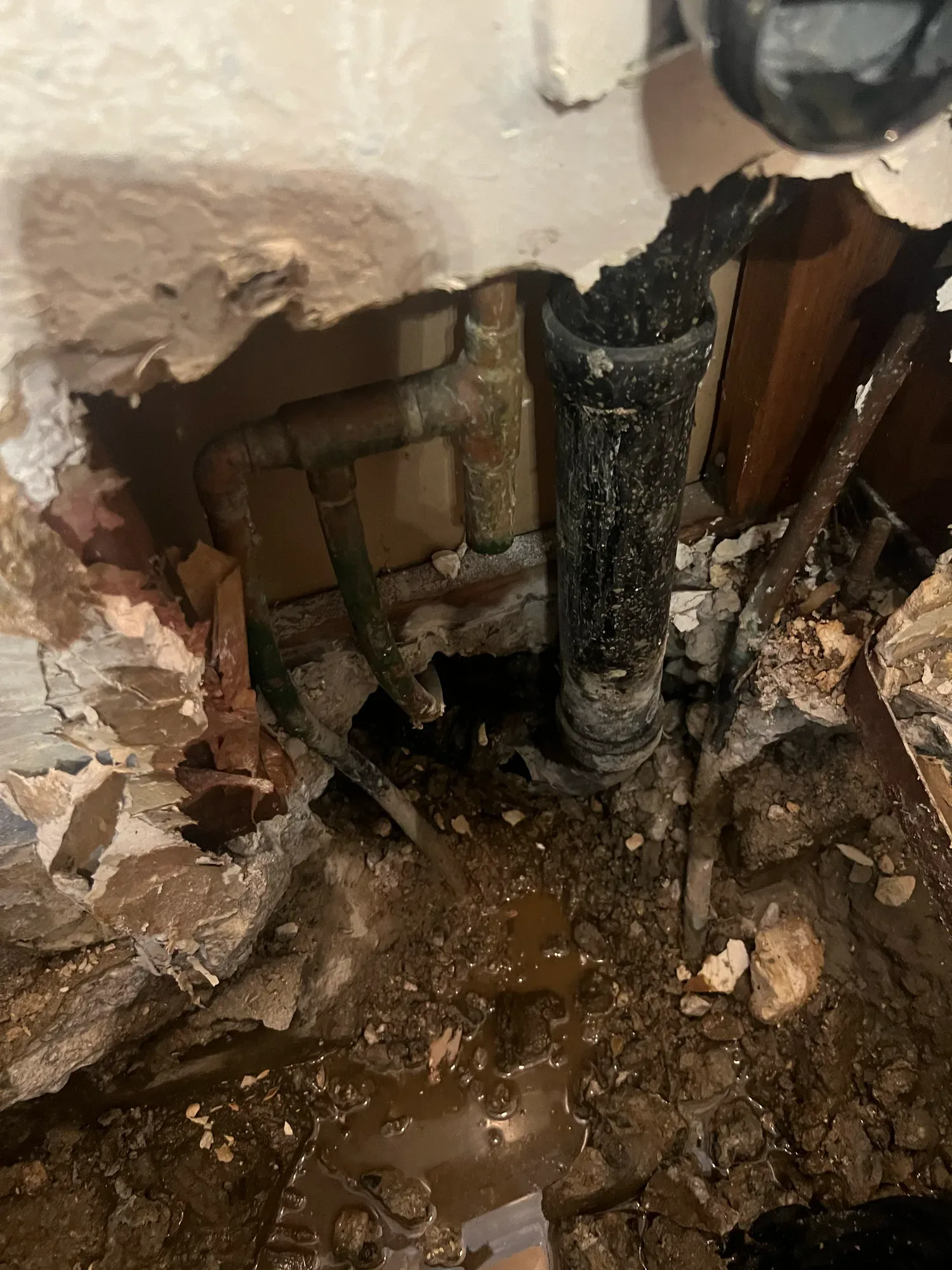 Opened wall and floor exposing old water pipes and a black drain pipe, with standing water in the dirt below