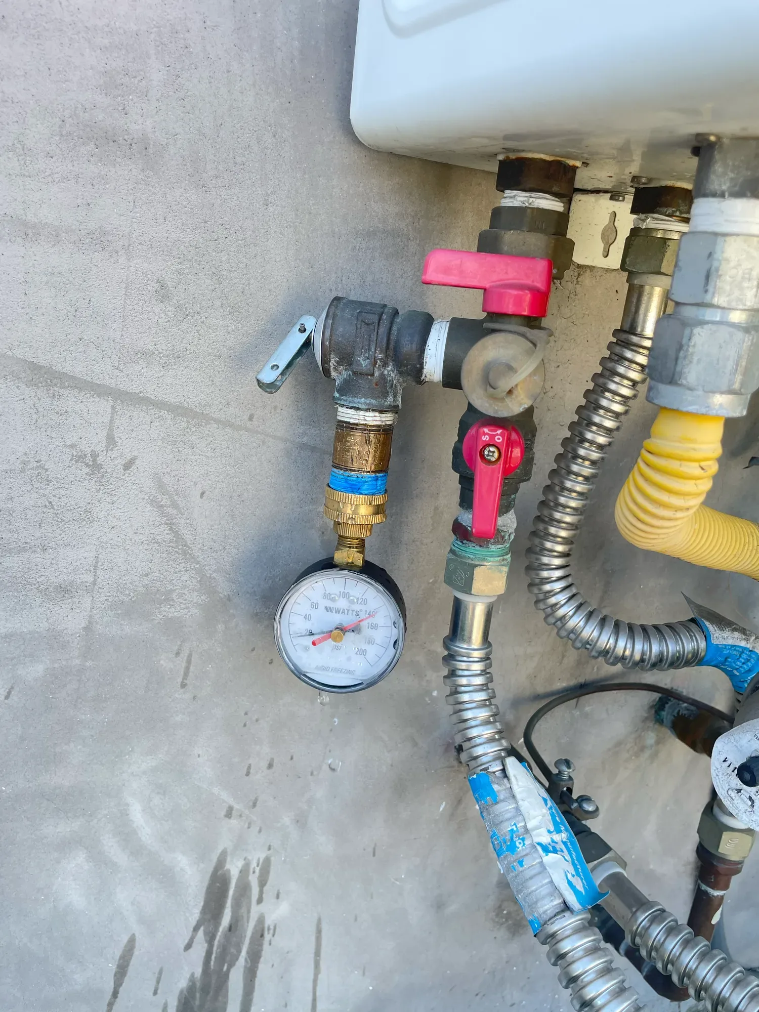 Pressure gauge fitted to the valves below a tankless water heater, with a yellow gas line and flexible water lines