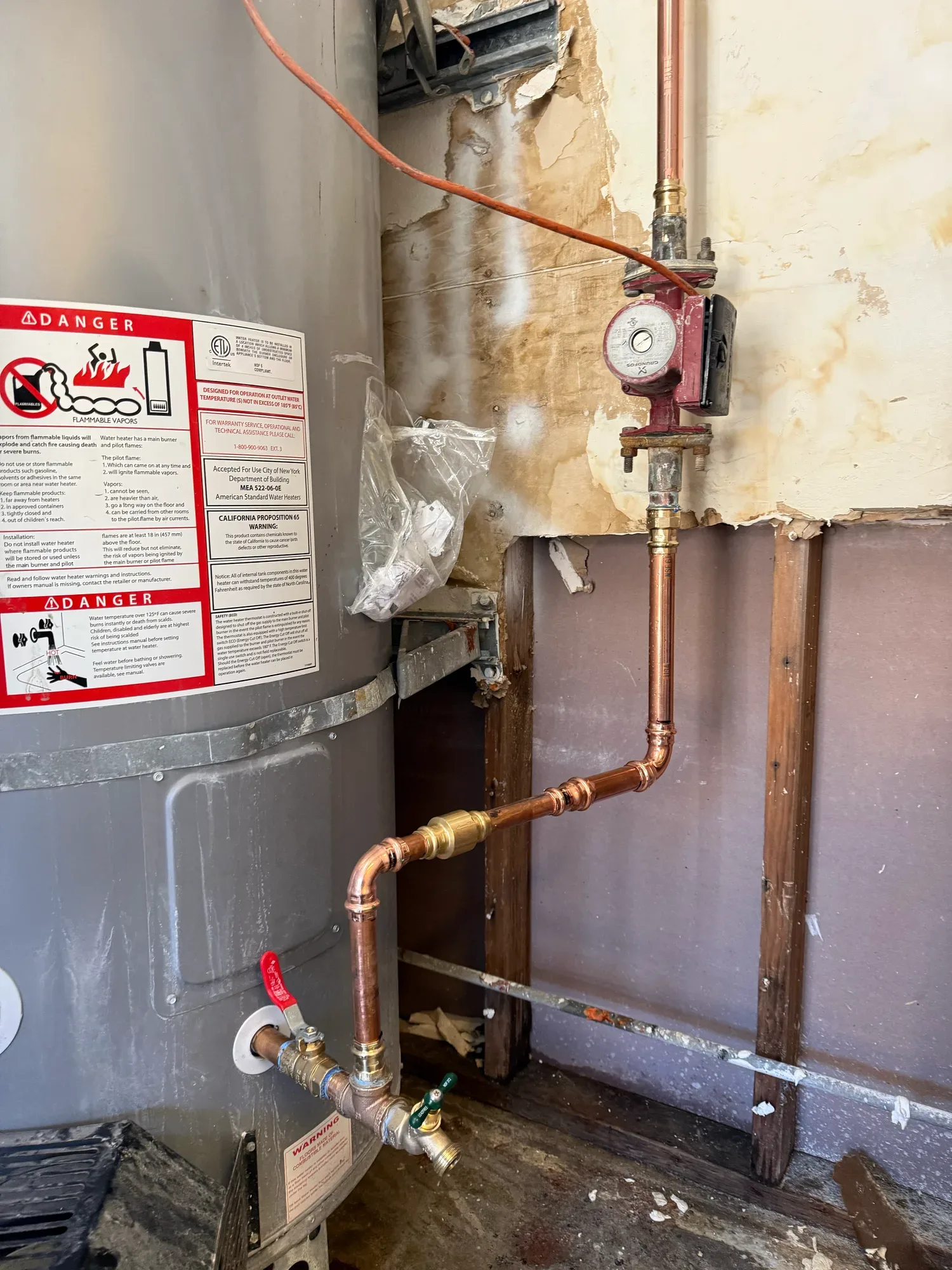 Tank water heater with a red circulation pump on the wall above it and new copper piping run down to its base