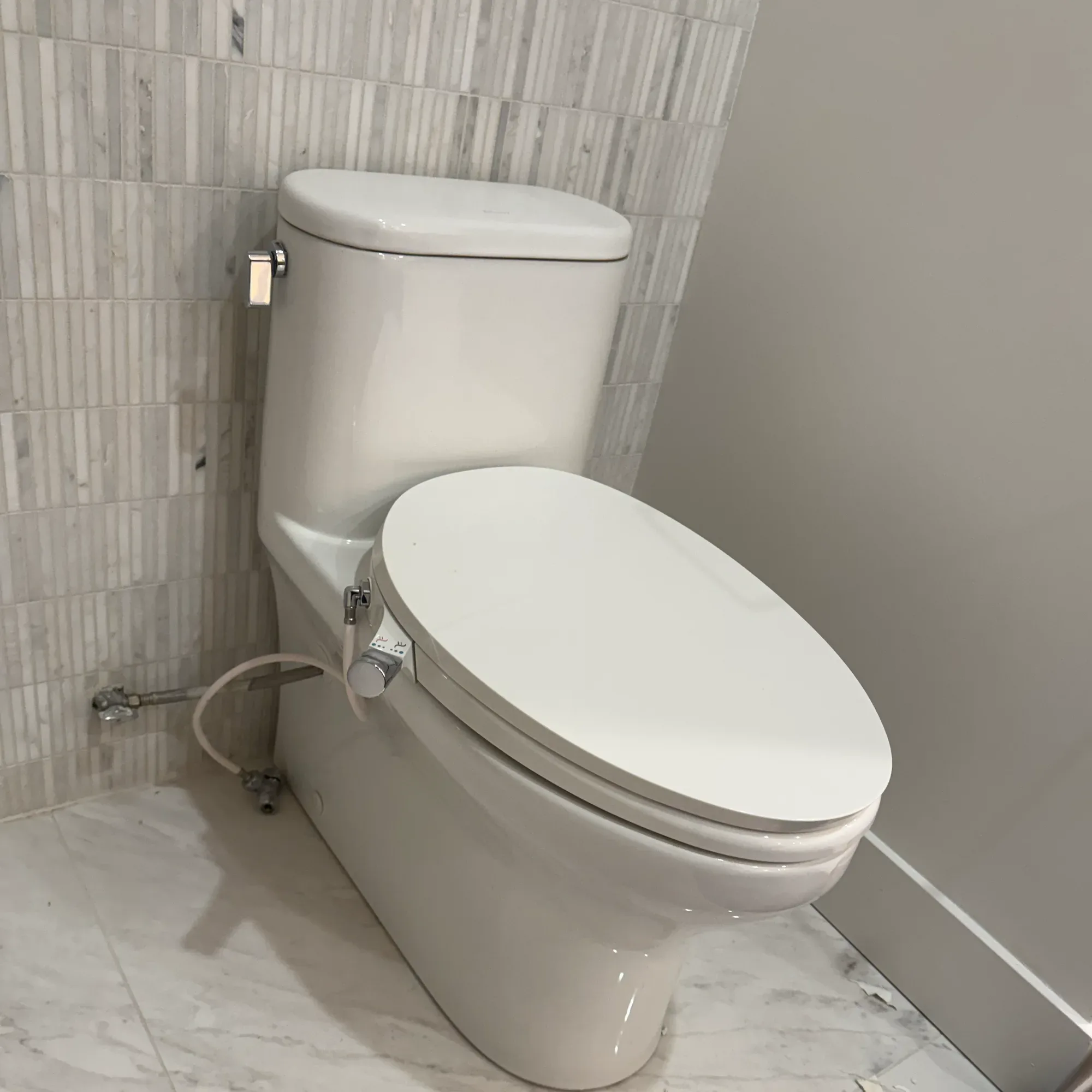 White toilet with a bidet seat and its supply hose connected, on a marble tile floor