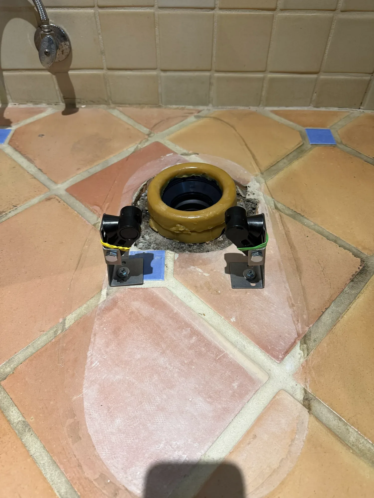 Open toilet drain with a new wax ring on the flange and two mounting brackets on a terracotta tile floor